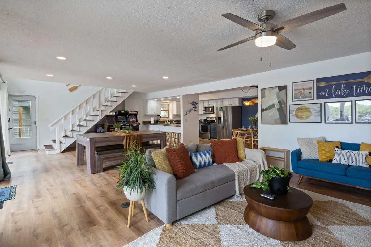 A spacious and comfortable open-plan living area featuring a sofa with colorful cushions, a wooden coffee table, a visible staircase leading to an upper level, and a kitchen area in the background. Natural light streams through large windows, enhancing the atmosphere and providing glimpses of the outdoors. Artwork on the walls adds to the cozy, welcoming vibe.