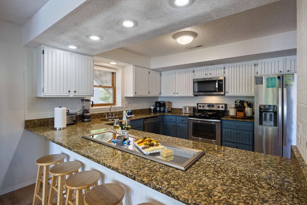 A welcoming kitchen scene with granite countertops, stainless steel appliances, wooden stools at the island, and a decorative platter with food and drinks. The layout encourages both cooking and social interaction, making it easy to host family breakfasts or entertain guests. Soft, inviting lighting enhances the room’s welcoming mood.