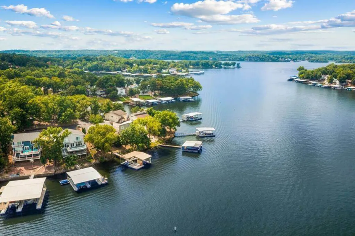 The image presents a sweeping view of the lake, with boathouses, docks, and surrounding trees lining the shore. The calm water glistens under the soft colors of sunset, setting a serene and inviting scene.