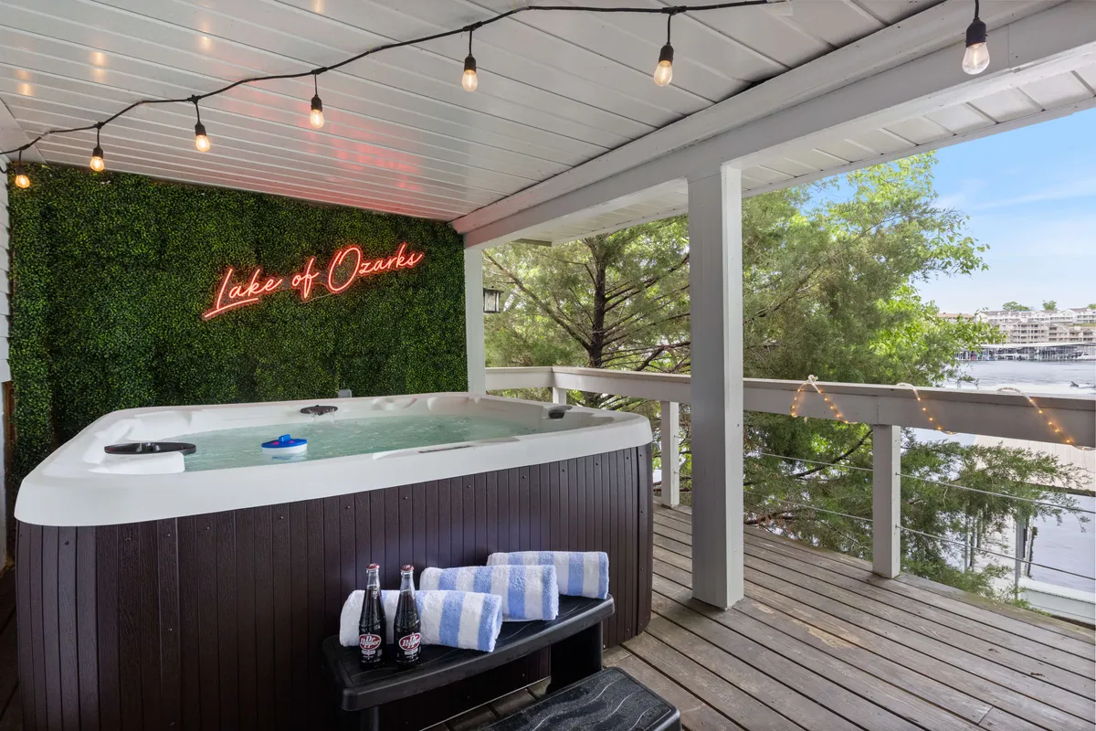 A close-up scene capturing a hot tub on the wooden deck, surrounded by towels, bottles of soda, and glowing with a neon sign. The lake is visible in the background, and the mood is leisurely, suggesting stargazing, relaxing with friends, or winding down after a day on the water.