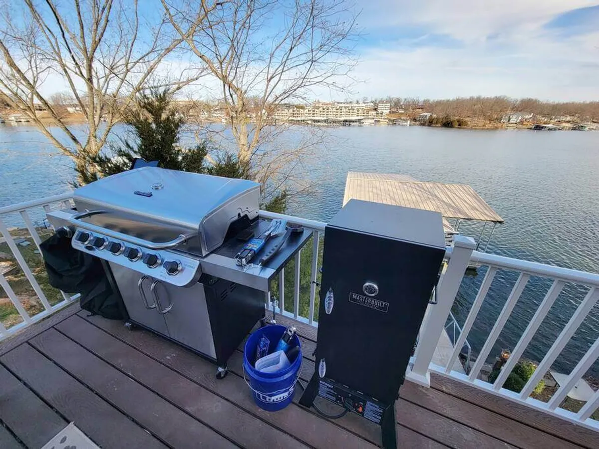This scene showcases an outdoor grill and smoker set against a beautiful water view. The setup suggests festive meals and casual gatherings, with ample space for outdoor dining and barbecues by the lake.