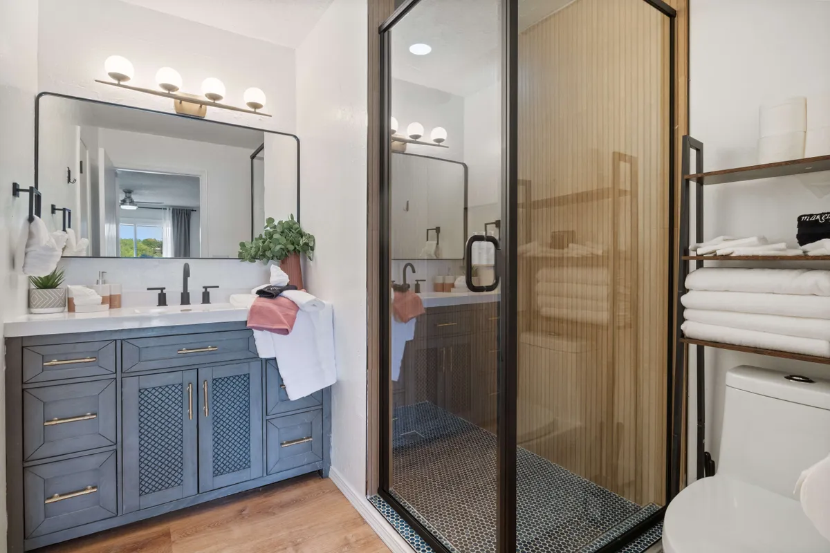 A bright, modern bathroom with a dual-sink vanity, large mirror, glass shower enclosure, and open shelving stocked with towels. The inviting design suggests plenty of space for morning routines or relaxing baths.