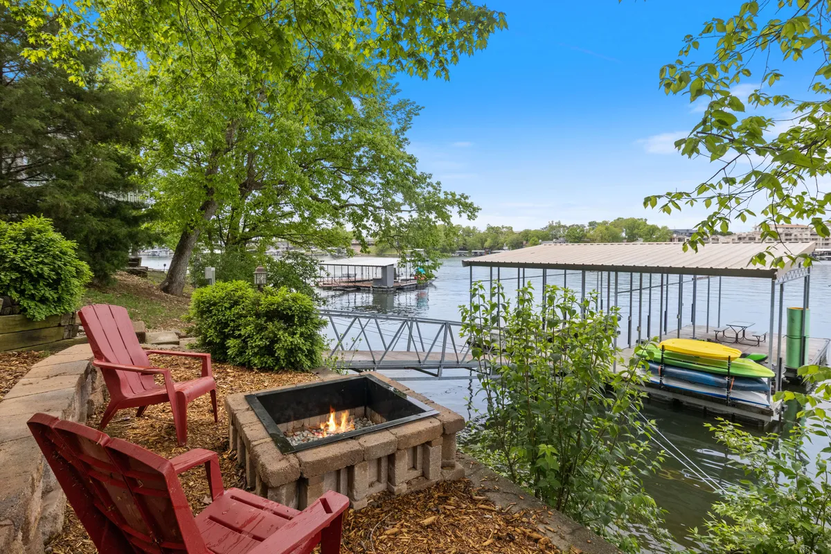 A set of adirondack chairs circles a fire pit on a grassy area near a dock and kayaks, with lush greenery and calm water in the background. The scene suggests gatherings, relaxation, and easy access to the water for boating or fishing.