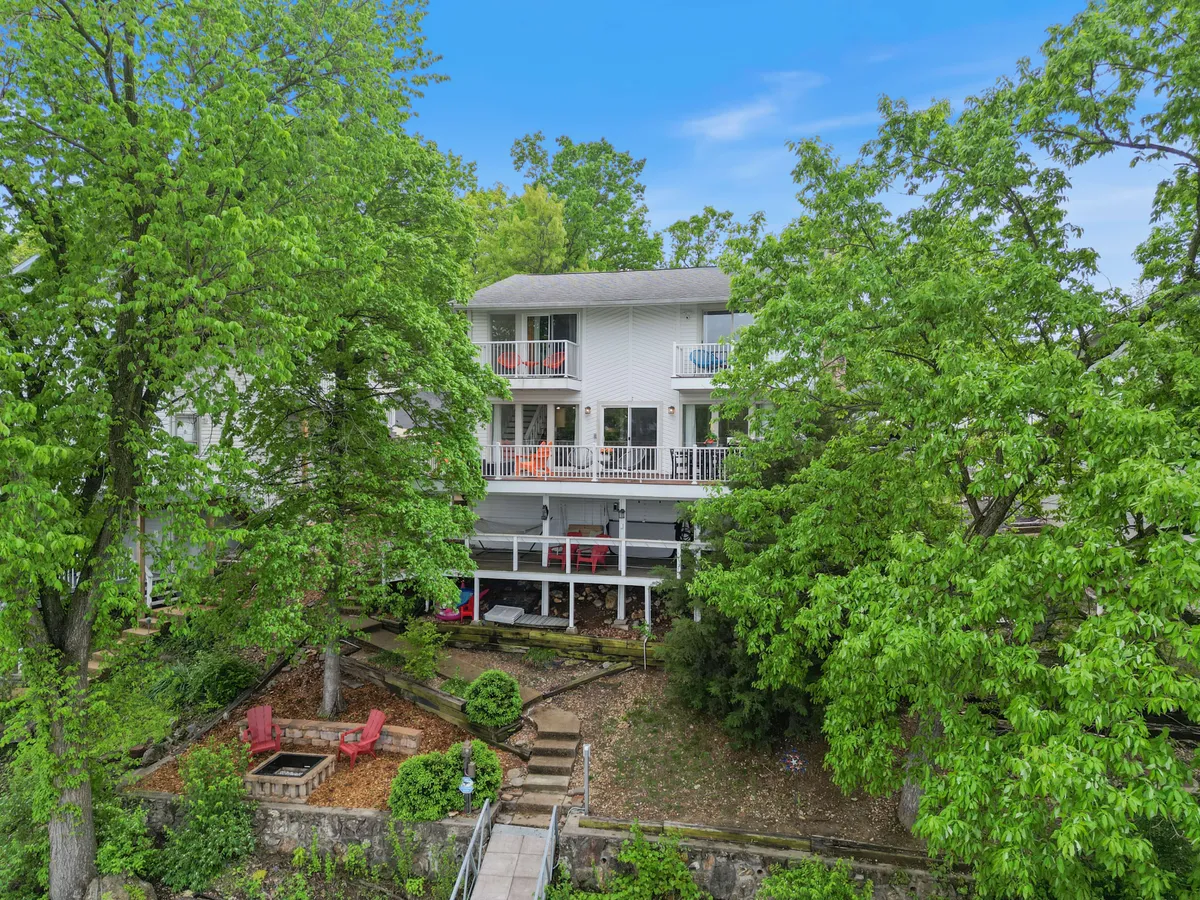 The image shows a multi-level house surrounded by lush green trees, a landscaped garden, and an inviting outdoor seating area. The setting is serene and welcoming, suggesting the perfect spot for outdoor relaxation and enjoying nature throughout the seasons.