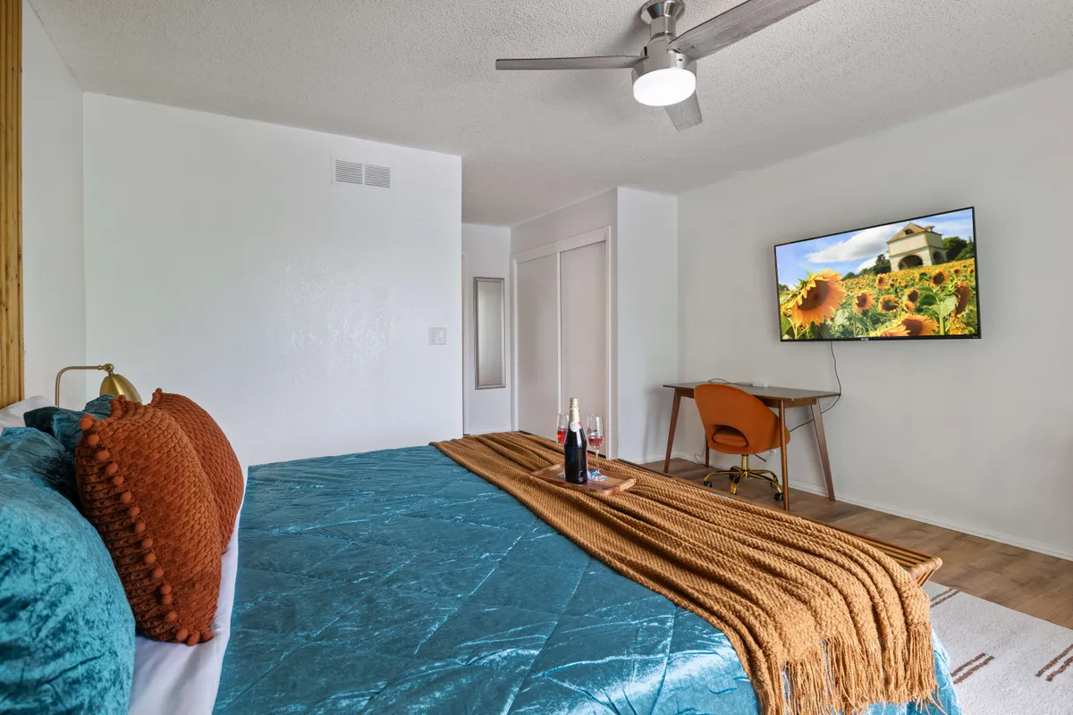 A cozy bedroom featuring a bed with teal bedding and orange pillows, a wall-mounted TV showing a sunflower field, and a wooden desk with a chair. The space feels inviting and functional, suitable for relaxing, watching TV, or working.